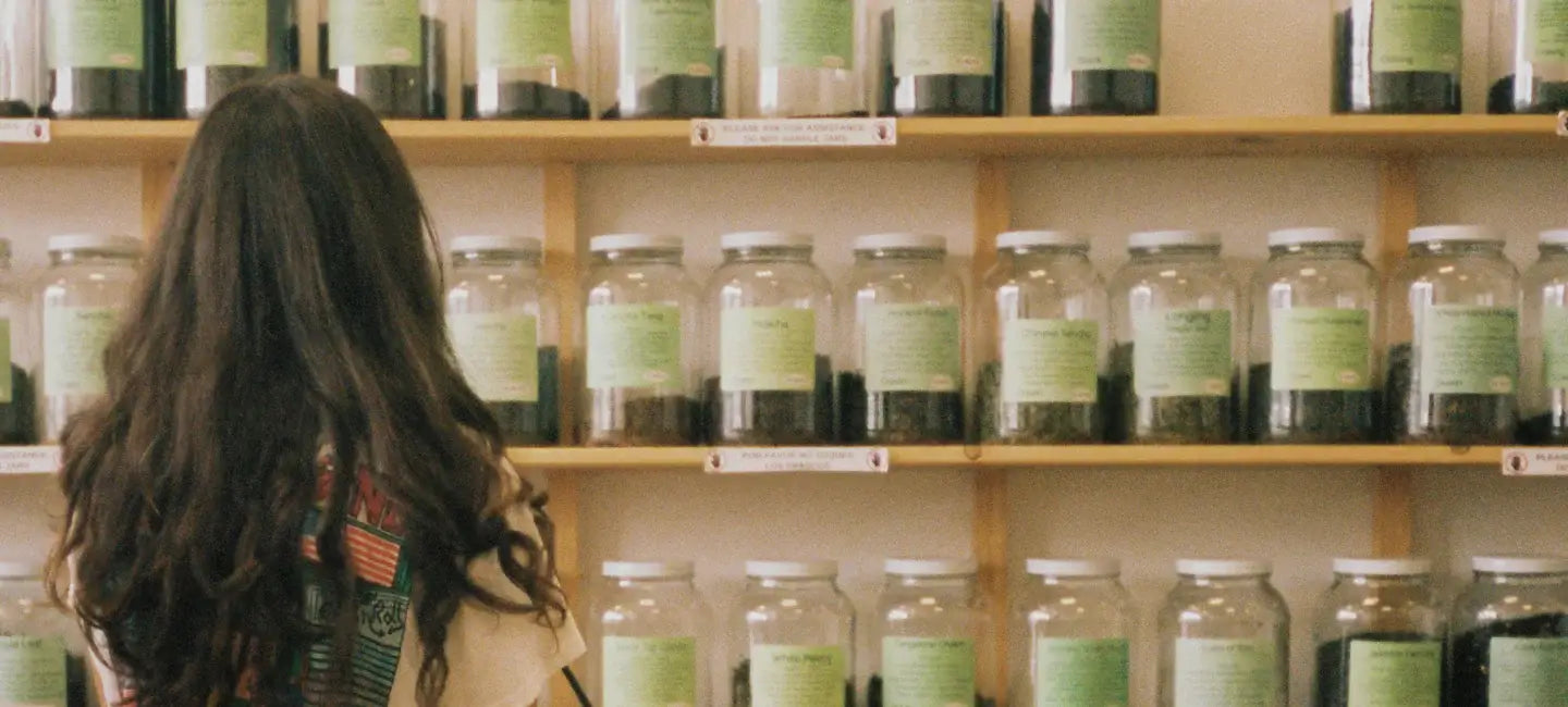 Woman with long dark hair browsing shelves of labeled glass herb jars in a warm apothecary-style shop